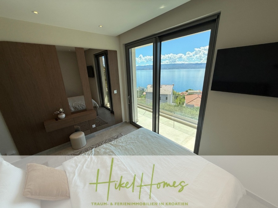 Erstbezug – ab sofort und Herbst genießen! Schlüsselfertige Meerblick Villa, 3SZ, Pool, … - Modernes Schlafzimmer mit großen Fenstern und Balkon mit Blick auf das Meer, mit einem Bett, einem Holzschrank, einem an der Wand montierten Fernseher und hellem Tageslicht. Der Text darüber lautet: "Hikel Homes Traum- & Ferienimmobilien in Kroatien. (44473)