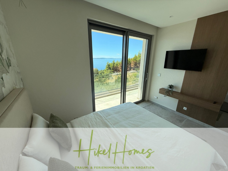 Erstbezug – ab sofort und Herbst genießen! Schlüsselfertige Meerblick Villa, 3SZ, Pool, … - Ein modernes Schlafzimmer mit einem großen Bett, einem an der Wand montierten Fernseher und Glasschiebetüren, die auf eine Terrasse mit Blick auf das Meer und die grüne Landschaft führen. Der Text am unteren Rand lautet "Hikel Homes Traum- & Ferienimmobilien in Kroatien". (44487)