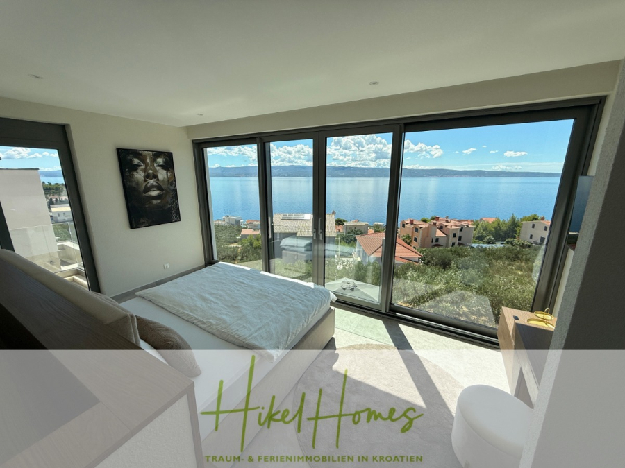 Erstbezug – ab sofort und Herbst genießen! Schlüsselfertige Meerblick Villa, 3SZ, Pool, … - Ein modernes Schlafzimmer mit großen, raumhohen Fenstern bietet einen Blick auf das Meer und die roten Dächer. Das Zimmer ist mit einem Bett, einem kleinen Schreibtisch und Wandkunst ausgestattet. Draußen sind Sonnenschein und blauer Himmel zu sehen. (44485)