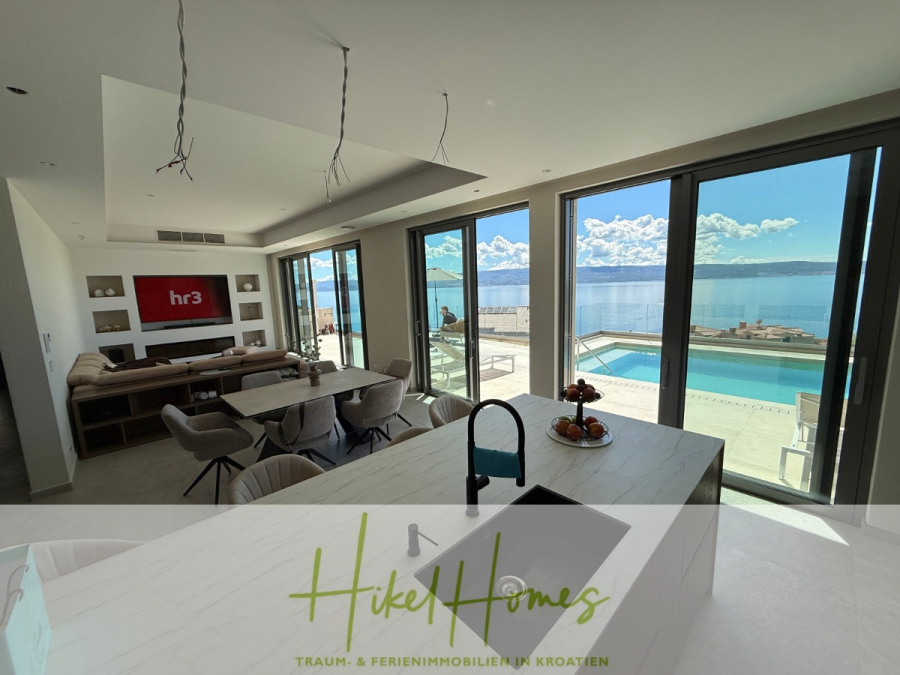 Erstbezug – ab sofort und Herbst genießen! Schlüsselfertige Meerblick Villa, 3SZ, Pool, … - Moderner offener Wohn- und Essbereich mit großen Glastüren, die den Blick auf die Terrasse, den Pool und die malerische Aussicht auf das Meer und die Berge unter blauem Himmel freigeben. Der Schriftzug "Hike Homes" ist am unteren Rand des Bildes zu sehen. (44474)