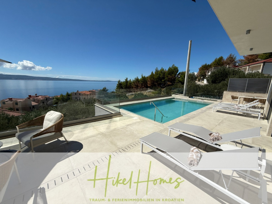 Erstbezug – ab sofort und Herbst genießen! Schlüsselfertige Meerblick Villa, 3SZ, Pool, … - Moderne Terrasse mit Liegestühlen, einem Pool und einem Glasgeländer mit Blick auf das Meer und die Häuser an der Küste, umgeben von Bäumen unter einem klaren blauen Himmel. Das Logo von "Hikel Homes" erscheint am unteren Rand. (44472)