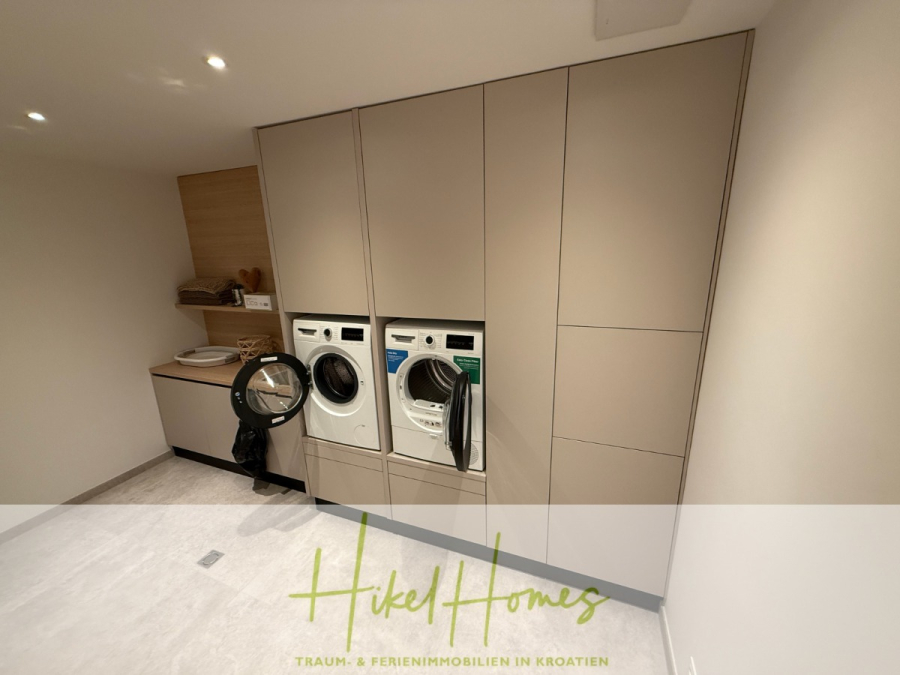 Erstbezug – ab sofort und Herbst genießen! Schlüsselfertige Meerblick Villa, 3SZ, Pool, … - Moderne Waschküche mit beigen Schränken, einer Waschmaschine und einem Trockner. Offene Regale nehmen Wäschevorräte auf. Der Raum ist minimalistisch eingerichtet und wirkt sauber und geordnet. Der Schriftzug "Hikel Homes" ist am Boden sichtbar. (44492)