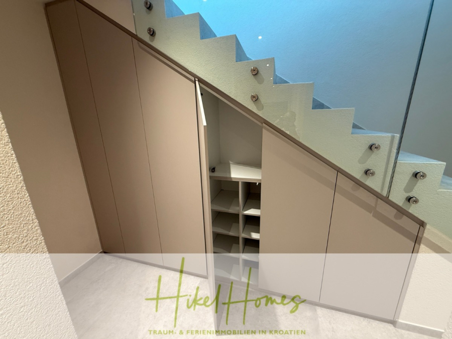 Erstbezug – ab sofort und Herbst genießen! Schlüsselfertige Meerblick Villa, 3SZ, Pool, … - Ein moderner Aufbewahrungsschrank mit offenen Regalen unter einer Treppe mit Glasgeländer. Die Schranktüren sind beige, und einige sind offen und geben den Blick auf geordnete Regale im Inneren frei. Das Logo "Hikel Homes" ist am unteren Rand eingeblendet. (44490)