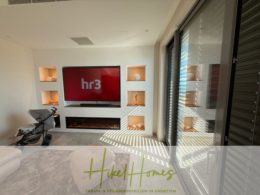 Erstbezug – ab sofort und Herbst genießen! Schlüsselfertige Meerblick Villa, 3SZ, Pool, … - Ein modernes Wohnzimmer mit einem großen Fernseher, der "hr3" zeigt, eingebauten Regalen mit Dekoration, einem elektrischen Kamin, einem Kinderwagen und Sonnenlicht, das durch teilweise geschlossene Jalousien fällt. Das Logo "Hikel Homes" überlagert den Boden. (44475)