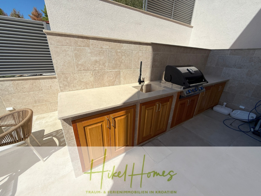 Erstbezug – ab sofort und Herbst genießen! Schlüsselfertige Meerblick Villa, 3SZ, Pool, … - Außenküche mit Spüle, Grill und Holzschränken vor einer beige gefliesten Wand. Auf der linken Seite ist ein Korbstuhl teilweise sichtbar. Das Sonnenlicht wirft Schatten auf den sauberen, modernen Raum. (44478)