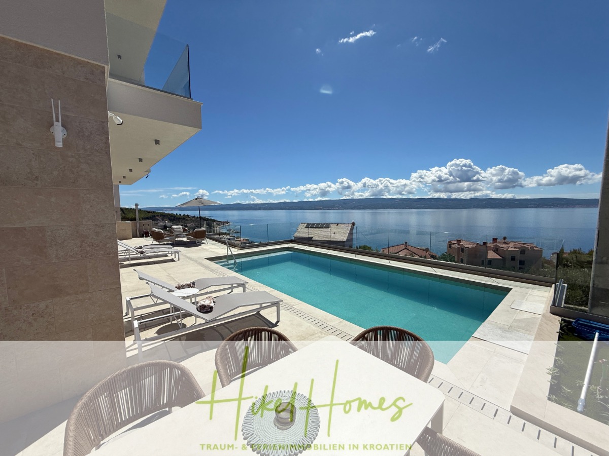 Moderne Terrasse mit Pool mit Blick auf das Meer, Sonnenliegen und Esstisch unter blauem Himmel. In der Ferne sind Wohnhäuser und Berge zu sehen. Der Text am unteren Rand lautet "Hike Homes". - Erstbezug – ab sofort und Herbst genießen! Schlüsselfertige Meerblick Villa, 3SZ, Pool, …