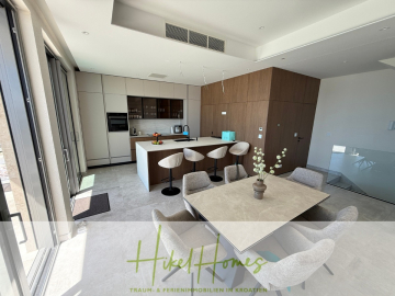 Moderne Küche und Essbereich mit hellen Schränken, einer zentralen Insel mit Barhockern und einem Esstisch mit gepolsterten Stühlen. Große Fenster sorgen für natürliches Licht. Das Branding lautet "Hikel Homes - Traum- & Ferienimmobilien in Kroatien". - Erstbezug – ab sofort und Herbst genießen! Schlüsselfertige Meerblick Villa, 3SZ, Pool, …