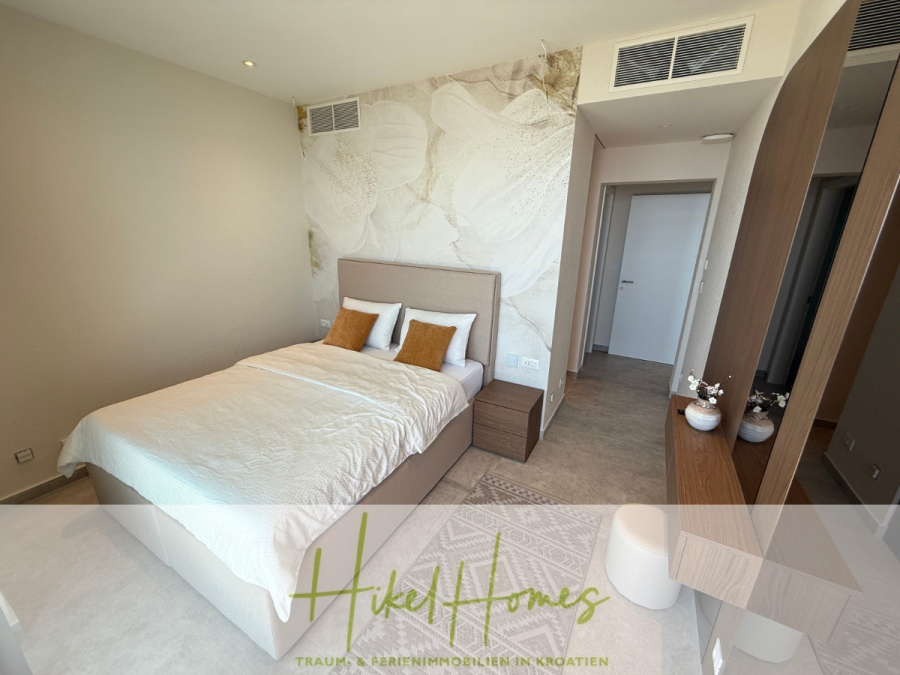Erstbezug – ab sofort und Herbst genießen! Schlüsselfertige Meerblick Villa, 3SZ, Pool, … - Modernes Schlafzimmer mit einem Doppelbett, weißer und beiger Bettwäsche, zwei braunen Akzentkissen, einer Wand mit abstrakter Kunst, einem hölzernen Kleiderschrank mit Spiegel und einem durch eine offene Tür einsehbaren Flur. (44481)