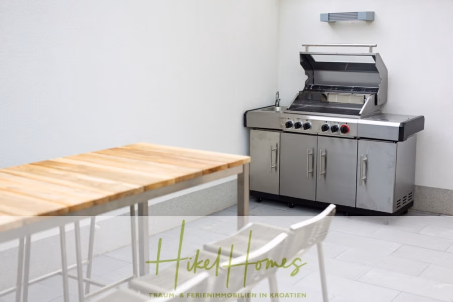 Erstbezug-Neubau Villa / Luxus Ausstattung / Pool / … - Außenterrasse mit Edelstahlgrill auf gefliestem Boden. Daneben stehen ein Holztisch und weiße Stühle. Das Ambiente besticht durch klare, minimalistische Linien und eine helle, neutrale Farbpalette. Das Logo unten lautet „Hikel Homes“. (42550)