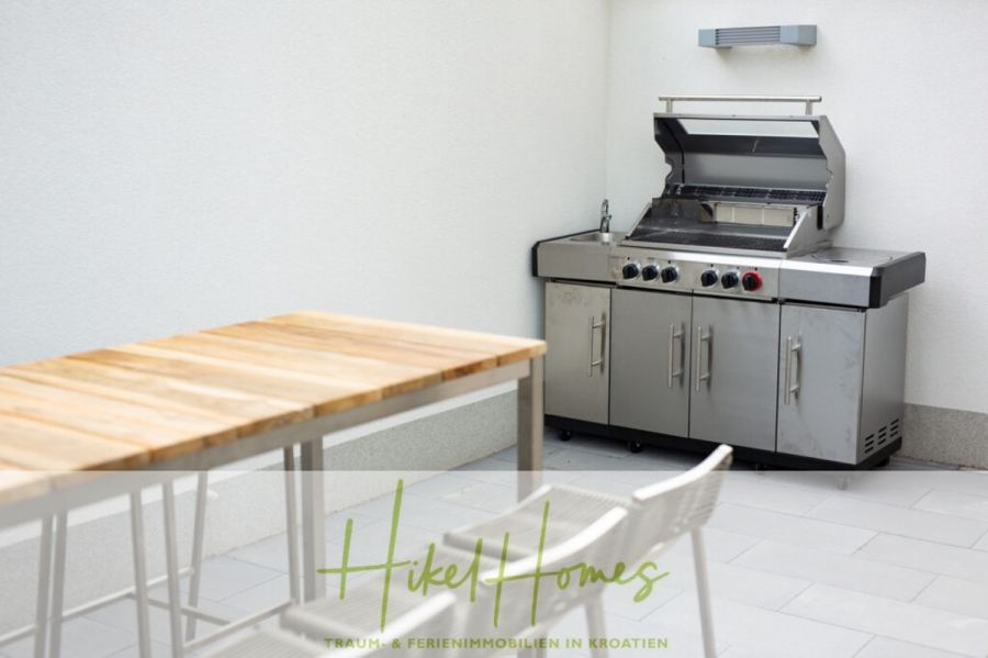 Erstbezug-Neubau Villa / Luxus Ausstattung / Pool / … - Eine moderne Außenterrasse mit einem Gasgrill aus Edelstahl, einem Holztisch und einem weißen Stuhl. Der Bereich ist sauber und minimalistisch mit hellen Fliesenböden und weißen Wänden. (45938)