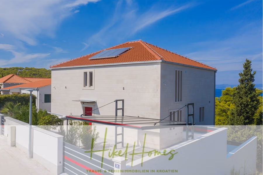Erstbezug-Neubau Villa / Luxus Ausstattung / Pool / … - Modernes zweistöckiges Haus mit rotem Ziegeldach und Solarmodulen, umgeben von einer weißen Wand. Üppiges Grün und strahlend blauer Himmel im Hintergrund. Unten befindet sich der Text „Hike Homes – Traumhafte Ferienimmobilien in Kroatien“. (42533)