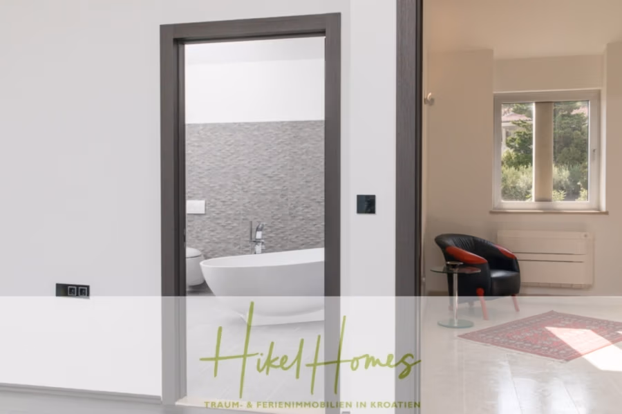 Erstbezug-Neubau Villa / Luxus Ausstattung / Pool / … - Ein modernes Interieur mit einem Badezimmer mit freistehender weißer Badewanne, die durch eine Tür sichtbar ist. Im angrenzenden Raum stehen ein rot-schwarzer Stuhl und ein kleiner Tisch neben einem Fenster mit viel Grün. Unten ist der Markentext „Hikel Homes“ eingeblendet. (42561)