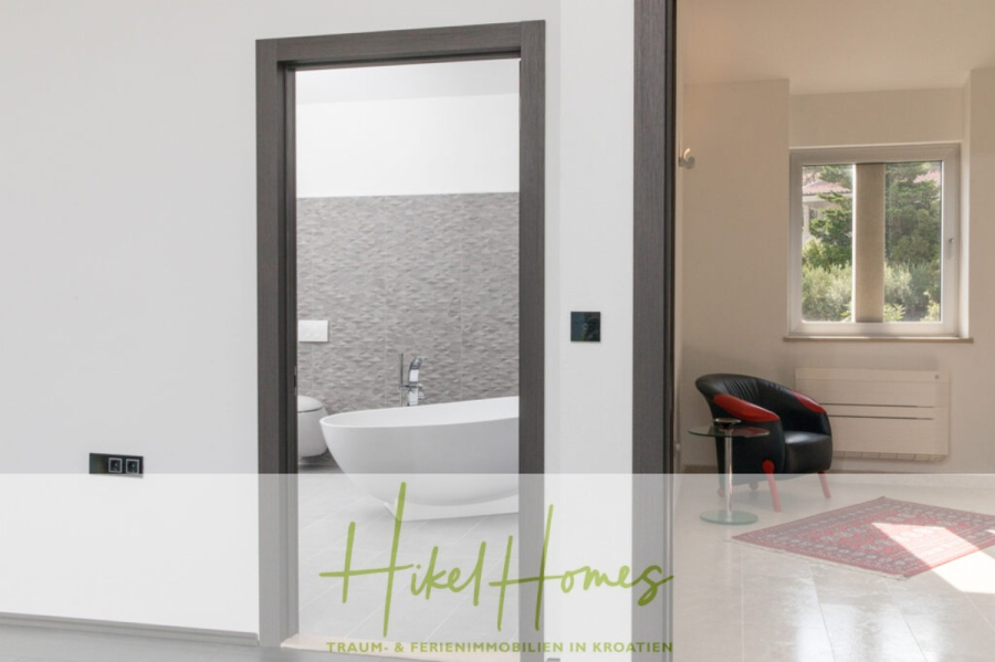 Erstbezug-Neubau Villa / Luxus Ausstattung / Pool / … - Ein modernes, minimalistisches Badezimmer mit einer weißen, freistehenden Badewanne und einer grauen, strukturierten Wand, von einem hellen Raum aus gesehen, mit einem schwarzen Stuhl, einem roten Kissen und einem Fenster, das Grün nach draußen zeigt. (45950)