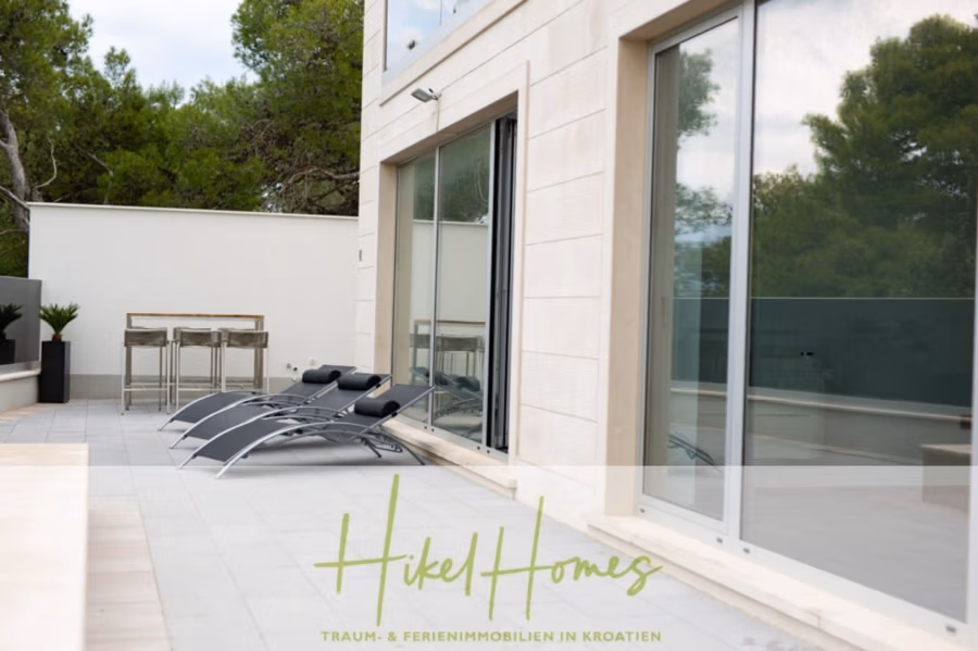 Erstbezug-Neubau Villa / Luxus Ausstattung / Pool / … - Moderner Terrassenbereich mit drei schwarzen Loungesesseln neben großen Glastüren eines beigen Gebäudes. Im Hintergrund steht ein hoher Bartisch mit Hockern, umgeben von Grün. (42540)