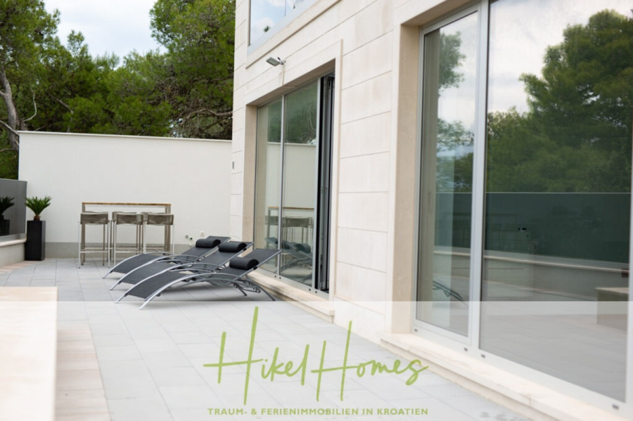 Erstbezug-Neubau Villa / Luxus Ausstattung / Pool / … - Moderne Terrasse mit drei schwarzen Loungesesseln vor großen Glastüren, daneben ein hoher Tisch mit Hockern, umrahmt von Bäumen. Der Text auf dem Bild lautet "Hikel Homes Traum- & Ferienimmobilien in Kroatien. (45927)