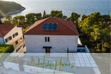 Ein modernes weißes Haus mit Sonnenkollektoren auf dem Dach, umgeben von Bäumen und benachbarten Häusern mit roten Dächern, mit Blick auf das blaue Meer in Kroatien. Das Logo "Hikel Homes" und deutscher Text erscheinen am unteren Rand. - Erstbezug-Neubau Villa / Luxus Ausstattung / Pool / …