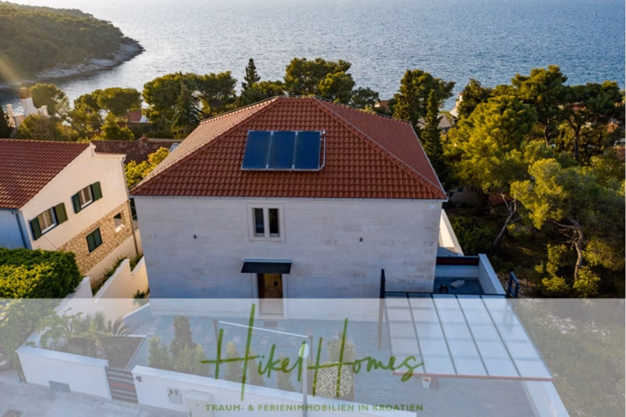 Erstbezug-Neubau Villa / Luxus Ausstattung / Pool / … - Luftaufnahme eines Hauses mit Solarmodulen auf dem Dach, umgeben von Bäumen und mit Blick auf ein Gewässer. Auch benachbarte Häuser haben rote Dächer. Das Bild trägt den Text „Hikel Homes“ und „Traum- & Ferienimmobilien in Kroatien“. (42531)