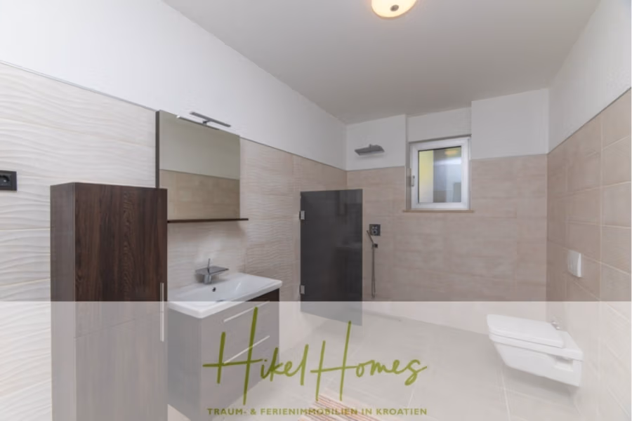 Erstbezug-Neubau Villa / Luxus Ausstattung / Pool / … - Modernes Badezimmer mit beigefarbenen Fliesen, einem Wandwaschbecken mit quadratischem Spiegel darüber, einem Holzschrank daneben, einer verglasten Dusche und einem Wand-WC. Über der Dusche befindet sich ein kleines Fenster. Die Textüberlagerung lautet „Hikel Homes“. (42556)
