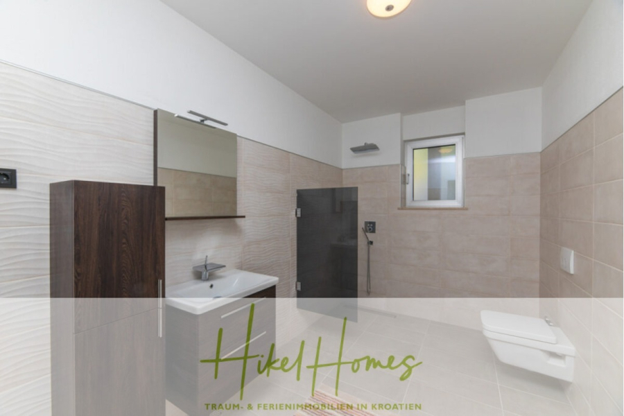 Erstbezug-Neubau Villa / Luxus Ausstattung / Pool / … - Geräumiges, modernes Badezimmer mit beigen Fliesen, einer begehbaren Dusche, einem an der Wand befestigten Waschbecken, einem Schrank aus dunklem Holz, einem großen Spiegel und einer an der Wand befestigten Toilette. Ein kleines Fenster lässt natürliches Licht herein. Das "Hikel Homes"-Logo ist am unteren Rand eingeblendet. (45945)