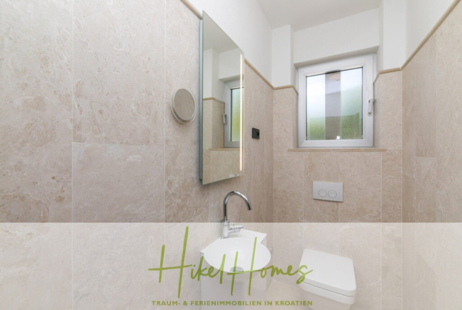 Erstbezug-Neubau Villa / Luxus Ausstattung / Pool / … - Ein kleines, modernes Badezimmer mit beigen Marmorwänden, einem quadratischen weißen Waschbecken, einem an der Wand montierten Wasserhahn, einem Spiegel und einem Milchglasfenster über einer Toilette mit verdecktem Spülkasten. Das Logo "Hikel Homes" ist am unteren Rand eingeblendet. (45953)