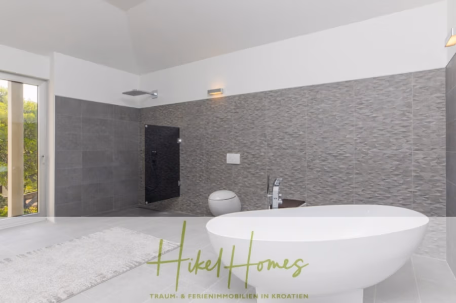 Erstbezug-Neubau Villa / Luxus Ausstattung / Pool / … - Modernes Badezimmer mit freistehender weißer Badewanne, Wand-WC und schwarzer Regendusche. Die Wände sind mit strukturierten grauen Fliesen gestaltet. Ein Fenster links sorgt für Tageslicht. Der Logotext unten lautet: „Hike Homes“ mit zusätzlichem kleineren Text. (42562)