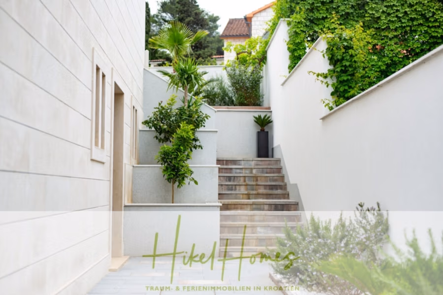 Erstbezug-Neubau Villa / Luxus Ausstattung / Pool / … - Eine moderne Außentreppe mit beigen Steinstufen führt hinauf, gesäumt von üppigem Grün und Topfpflanzen. Die Wände sind weiß, und in der Ferne ist ein Hausdach zu sehen. Unten steht der Text „Hikel Homes – Traum- & Ferienimmobilien in Kroatien“. (42552)