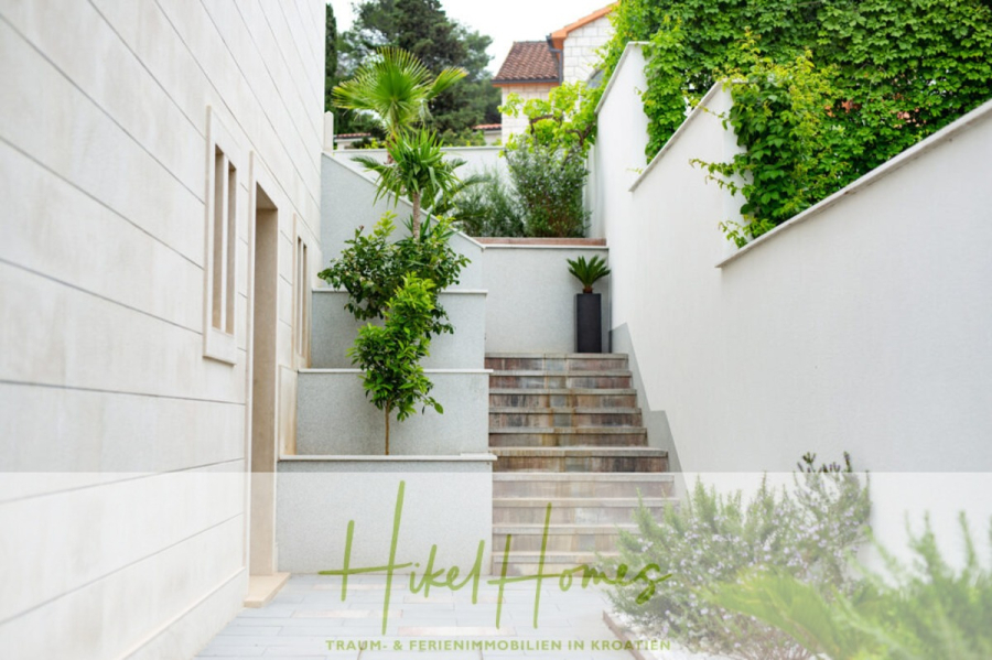 Erstbezug-Neubau Villa / Luxus Ausstattung / Pool / … - Eine Steintreppe im Freien mit modernen weißen Wänden, Topfpflanzen und Grünpflanzen führt zu einer sonnigen Terrasse. "Hikel Homes" und ein Text in deutscher Sprache sind auf einem durchsichtigen Banner am unteren Rand zu sehen. (45941)