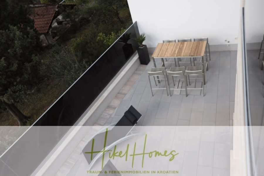 Erstbezug-Neubau Villa / Luxus Ausstattung / Pool / … - Die Draufsicht zeigt einen geräumigen Balkon mit hellgrauen Fliesen, einem Holztisch mit sechs Metallstühlen und einem einzelnen Loungesessel. Im Hintergrund sind üppiges Grün und ein Teil eines Hausdachs zu sehen. (42553)