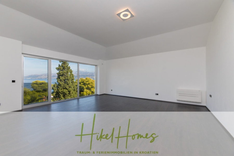 Erstbezug-Neubau Villa / Luxus Ausstattung / Pool / … - Ein geräumiger, leerer Raum mit großen Fenstern, die einen malerischen Blick auf das Meer und das Grün bieten, mit grauem Bodenbelag, weißen Wänden und einer modernen Deckenleuchte. Der Schriftzug "Hikel Homes" ist am unteren Rand eingeblendet. (45943)