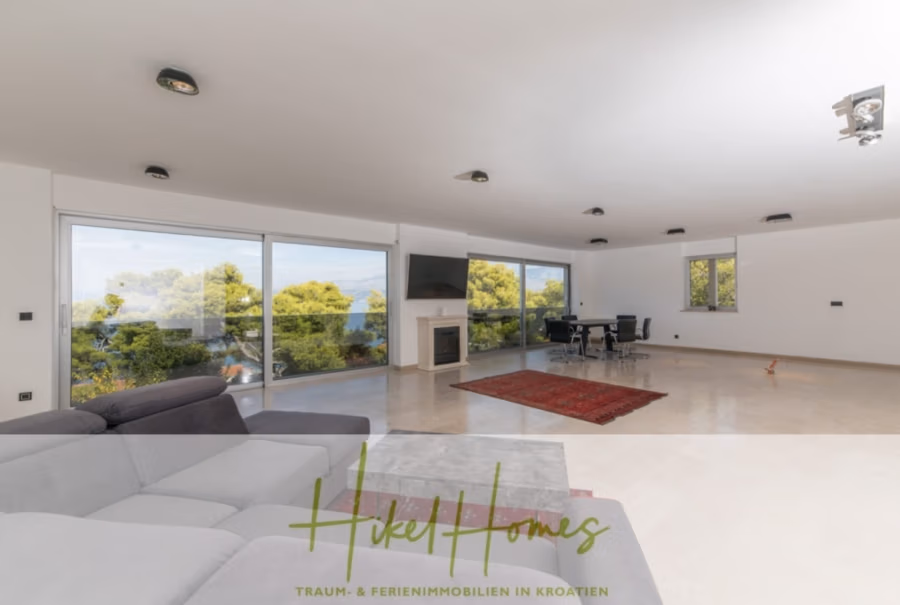 Erstbezug-Neubau Villa / Luxus Ausstattung / Pool / … - Geräumiges, modernes Wohnzimmer mit großen Fenstern und Blick auf die Bäume. Zur Ausstattung gehören ein graues Sofa, ein an der Wand montierter Fernseher über dem Kamin, ein roter Teppich und ein schwarzer Tisch mit Stühlen. Helles und luftiges Ambiente. (42543)