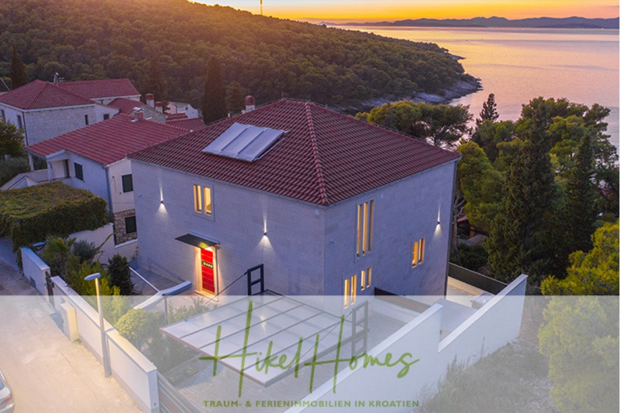 Erstbezug-Neubau Villa / Luxus Ausstattung / Pool / … - Modernes weißes Haus mit rotem Dach in der Nähe des Meeres bei Sonnenuntergang, umgeben von Bäumen. Warme Lichter leuchten aus den Fenstern. Das "Hikel Homes"-Logo und der deutsche Text erscheinen am unteren Rand des Bildes. (45916)