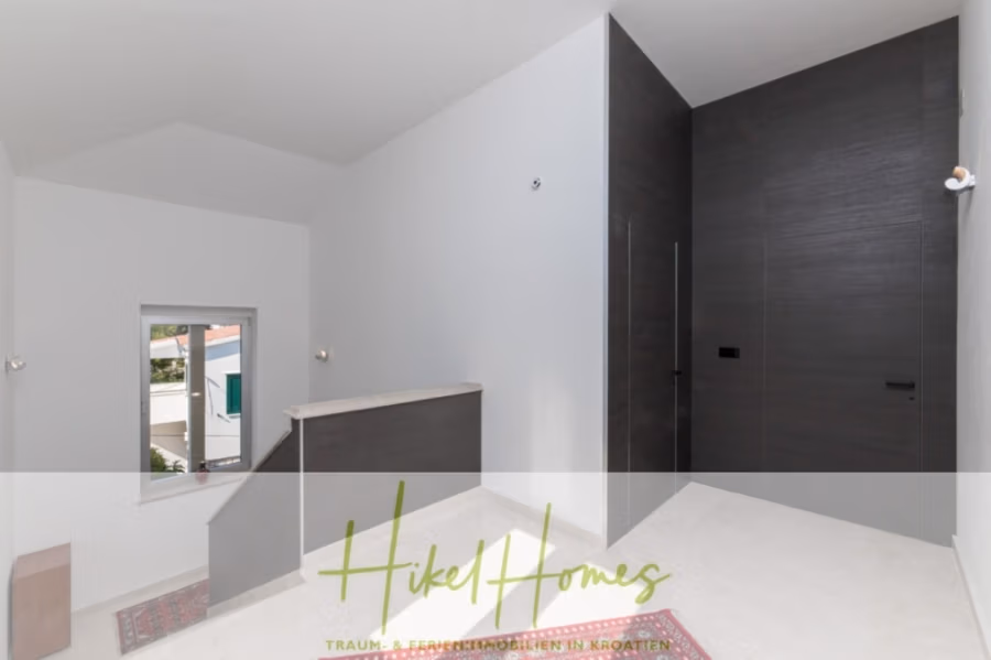 Erstbezug-Neubau Villa / Luxus Ausstattung / Pool / … - Minimalistisches Interieur mit weißen Wänden und einer schwarzen Akzentwand. Eine Treppe mit Holzgeländer und ein kleines Fenster sind sichtbar. Der Raum ist modern und klar gestaltet und besticht durch Fliesenboden und dezente Beleuchtung. Die Textüberlagerung lautet „Hikel Homes“. (42560)