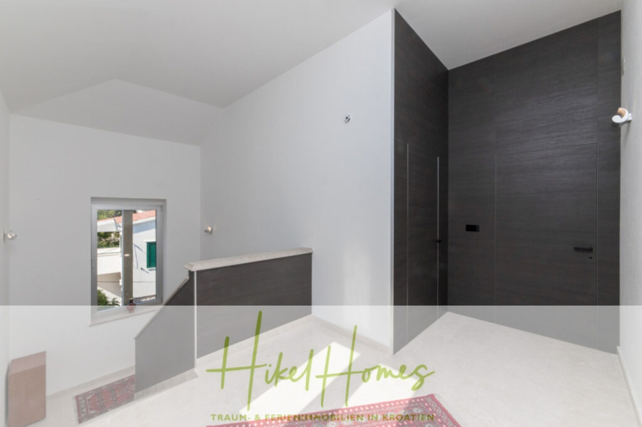 Erstbezug-Neubau Villa / Luxus Ausstattung / Pool / … - Ein heller, moderner Flur mit weißen Wänden, einem kleinen Fenster, dunklen Holztüren und einem hellen Boden mit einem gemusterten Teppich. Der Text "Hikel Homes" ist am unteren Rand des Bildes eingeblendet. (45949)