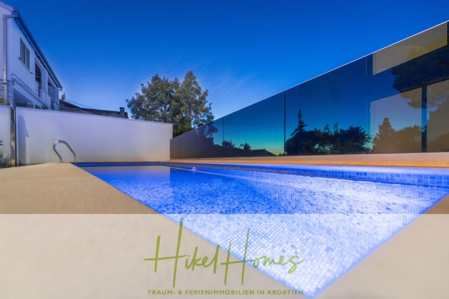 Erstbezug-Neubau Villa / Luxus Ausstattung / Pool / … - Modernes Freibad, das in der Abenddämmerung mit blauen Lichtern leuchtet, umgeben von einem Glaszaun, mit einem Haus und Bäumen im Hintergrund. Textüberlagerung: "Hikel Homes - Traum- & Ferienimmobilien in Kroatien. (45929)