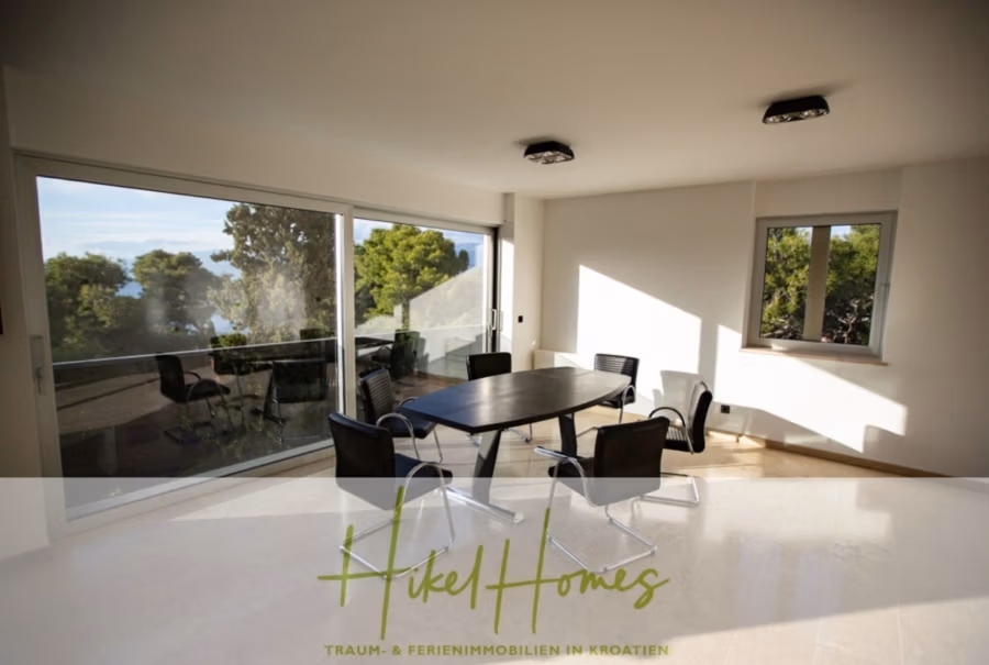 Erstbezug-Neubau Villa / Luxus Ausstattung / Pool / … - Moderner Essbereich mit dunklem ovalem Tisch und sechs schwarzen Stühlen, umgeben von großen Fenstern mit Blick ins Grüne und aufs Meer. Sonnenlicht strömt in den Raum und schafft eine helle und luftige Atmosphäre. Der Logotext überlagert den unteren Bildrand. (42548)