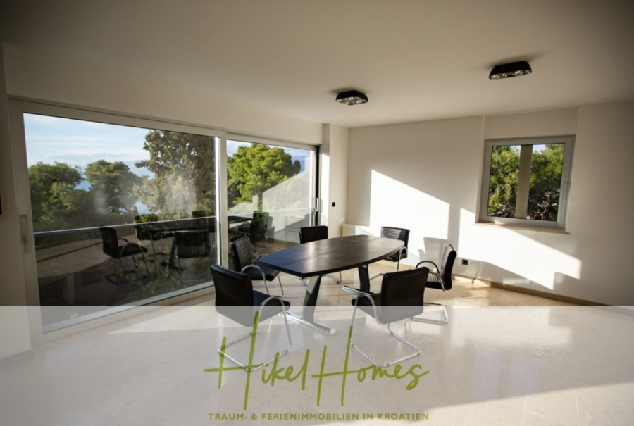 Erstbezug-Neubau Villa / Luxus Ausstattung / Pool / … - Ein moderner, sonnendurchfluteter Raum mit großen Fenstern und Glastüren, in dem ein schwarzer Tisch mit fünf schwarzen Stühlen steht. Draußen sind Grünflächen und ein entfernter Blick auf das Meer zu sehen. Der Text "Hikel Homes" erscheint am unteren Rand. (45937)