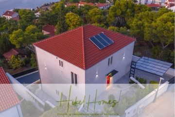 Luftaufnahme eines modernen weißen Hauses mit rotem Ziegeldach und Sonnenkollektoren, umgeben von Pinien und anderen Häusern in Küstennähe. Das Bild enthält das "Hike Homes"-Logo am unteren Rand. - Erstbezug-Neubau Villa / Luxus Ausstattung / Pool / …