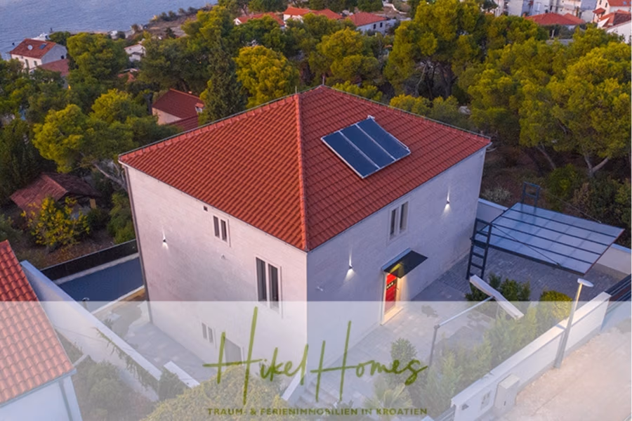 Erstbezug-Neubau Villa / Luxus Ausstattung / Pool / … - Luftaufnahme eines modernen, quadratischen Hauses mit rotem Ziegeldach und Solarmodulen, umgeben von Bäumen und Nachbarhäusern. Im Hintergrund ist ein Küstengewässer zu sehen. Das Bild verfügt über eine halbtransparente Überlagerung mit dem Schriftzug „Hike Homes“. (42532)