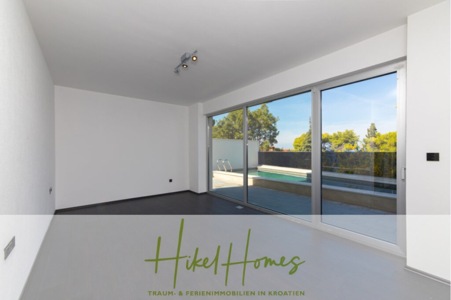 Erstbezug-Neubau Villa / Luxus Ausstattung / Pool / … - Helles, leeres, modernes Wohnzimmer mit großen Glasschiebetüren, die sich zu einer Terrasse und einem Pool hin öffnen, mit weißen Wänden, grauem Bodenbelag und Blick auf Bäume und den blauen Himmel. Der Schriftzug "Hikel Homes" erscheint am unteren Rand. (45925)