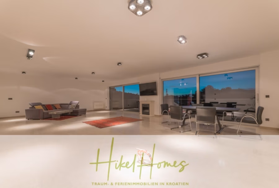 Erstbezug-Neubau Villa / Luxus Ausstattung / Pool / … - Ein geräumiges, modernes Wohnzimmer mit einem großen grauen Sofa, einem roten Teppich und einem Esstisch mit schwarzen Stühlen. Große Fenster bieten Blick auf eine Terrasse und Bäume. Der Raum wird von Deckenleuchten beleuchtet. Das Bild zeigt unten ein Logo und Text. (42547)