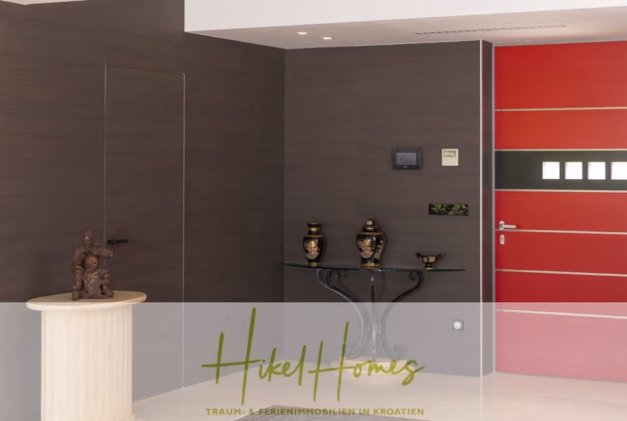 Erstbezug-Neubau Villa / Luxus Ausstattung / Pool / … - Modernes Interieur mit dunkler Holzwand, einem Glastisch mit dekorativen Vasen und einer eleganten rot-schwarzen Tür. Eine kleine Skulptur steht auf einem runden Sockel. Der Briefkopf lautet „Hikel Homes – Traum- & Ferienimmobilien in Kroatien“. (42545)