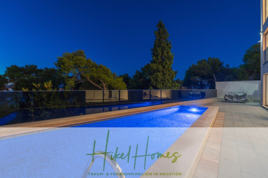 Erstbezug-Neubau Villa / Luxus Ausstattung / Pool / … - Ein moderner, nachts beleuchteter Außenpool, umgeben von Bäumen und einer gefliesten Terrasse, in deren Nähe ein überdachtes Auto geparkt ist. Der Schriftzug "Hikel Homes Traum- & Ferienimmobilien in Kroatien" erscheint quer über den Boden. (45930)