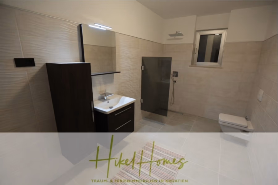 Erstbezug-Neubau Villa / Luxus Ausstattung / Pool / … - Modernes Badezimmer mit beigefarbenen Fliesen, einem dunklen Holzwaschtisch mit weißem Waschbecken, einem Wandspiegel und einer verglasten Dusche. Es gibt ein Fenster in Deckennähe und eine wandmontierte Toilette. Der Markenname lautet „Hikel Homes“. (42558)