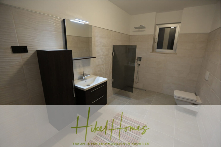 Erstbezug-Neubau Villa / Luxus Ausstattung / Pool / … - Modernes Badezimmer mit beigen Fliesen, einem Waschtisch aus dunklem Holz mit Spiegel, einem an der Wand befestigten Waschbecken, einer begehbaren Glasdusche, einem kleinen Fenster und einer an der Wand befestigten Toilette. Der Boden ist hell, und der Raum wirkt sauber und minimalistisch. (45947)