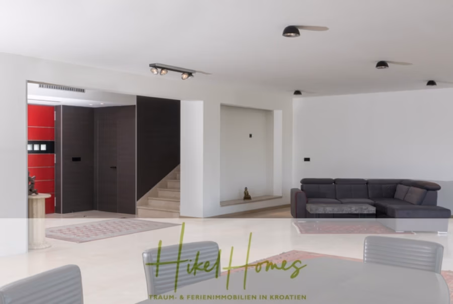 Erstbezug-Neubau Villa / Luxus Ausstattung / Pool / … - Geräumiges, modernes Wohnzimmer mit grauem Ecksofa, Glascouchtisch und minimalistischer Einrichtung. Der offene Grundriss führt zu einer Treppe. Weiße Wände und beigefarbener Boden verstärken das luftige Ambiente. Rote Akzente im Eingangsbereich. Deckenleuchten erhellen den Raum. (42546)