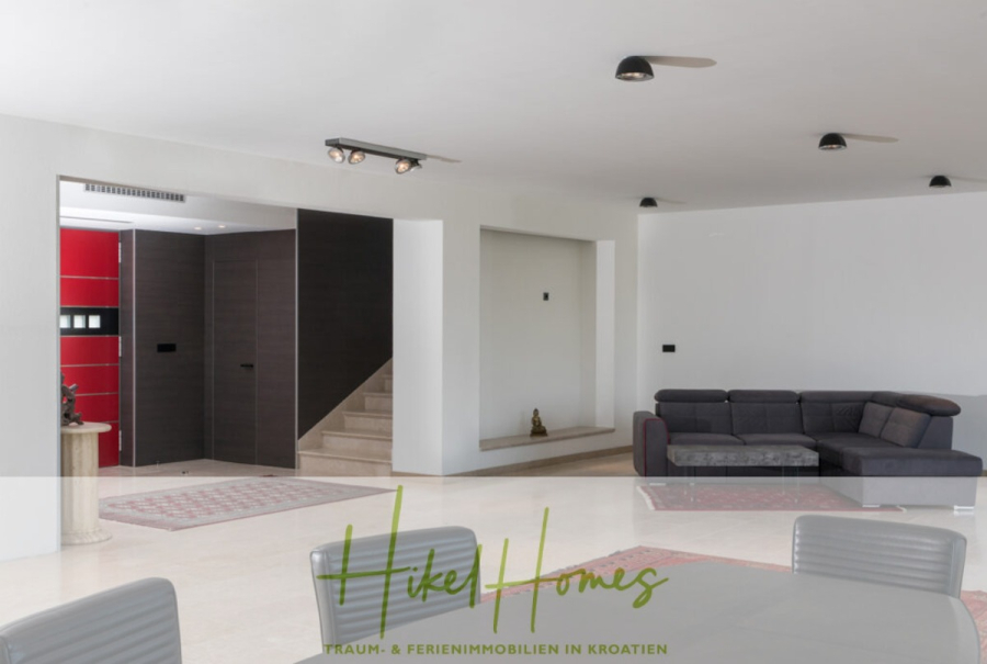 Erstbezug-Neubau Villa / Luxus Ausstattung / Pool / … - Modernes, minimalistisches Wohnzimmer mit weißen Wänden, einem grauen Sektionssofa, einem Couchtisch aus Glas und einem offenen Bereich, der zu einer Treppe mit roten und braunen Türen führt. Das Logo "Hike Homes" ist am unteren Rand eingeblendet. (45934)