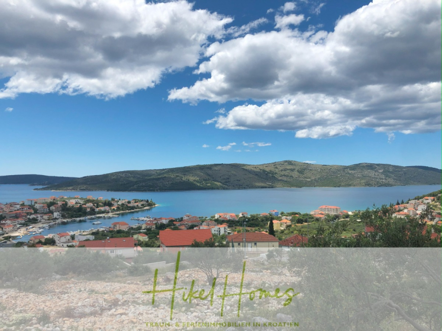 Meerblick – Schlüsselfertige Villa Princ in Seget Vranijca – modernste Technik – 3 SZ - Blick auf eine Küstenstadt mit roten Dächern, blaugrünem Wasser und üppigen Hügeln bei teilweise bewölktem Himmel. Im Vordergrund ist ein transparentes Banner mit dem Text "Hikel Homes" zu sehen. (45484)
