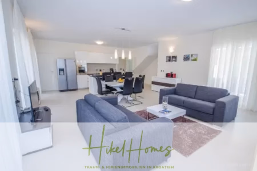 Villa im Mittelmeerstil mit 3 SZ, Pool, Dachterrasse mit Meerblick (2) - Modernes Wohnzimmer mit hellen Wänden, zwei grauen Sofas, einem weißen Couchtisch und einem Essbereich mit schwarzen Stühlen. Offene Küche mit Edelstahlgeräten. Große Fenster mit transparenten Vorhängen. Text-Overlay am unteren Rand. (42200)