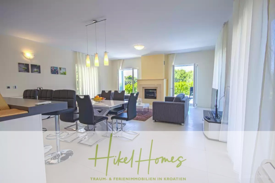 Villa im Mittelmeerstil mit 3 SZ, Pool, Dachterrasse mit Meerblick (2) - Moderner, heller, offener Wohn- und Essbereich mit großen Fenstern, einem Kamin, einem Esstisch, einem Sofa, einem Fernseher und Zugang zu einer begrünten Terrasse im Freien. Die Textüberlagerung lautet "Hikel Homes" in grün. (45796)