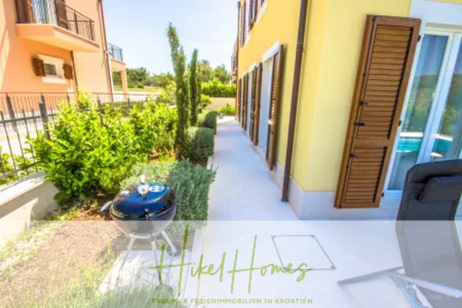 Villa im Mittelmeerstil mit 3 SZ, Pool, Dachterrasse mit Meerblick (2) - Ein sonniger Innenhof neben einem gelben Haus mit braunen Fensterläden, einem kleinen Garten, einem runden Grill und Sitzgelegenheiten im Freien. Grüne Sträucher säumen den Gehweg, und links ist ein weiteres Gebäude zu sehen. Der Text am unteren Rand lautet "Hikel Homes". (45783)